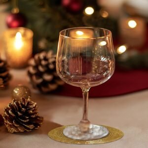 A festive table setting featuring a wine glass placed on a gold beaded coaster, surrounded by holiday decor including candles, pinecones, and ornaments.